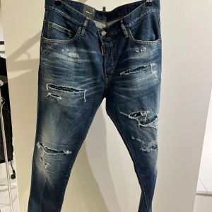 Blå slitna jeans - Säljer ett par helt nya blå Dsquared2 med coola slitningar och en avslappnad look. Perfekta för en casual stil och passar till nästan allt. De har en klassisk femficksdesign och knappar i gylfen.
