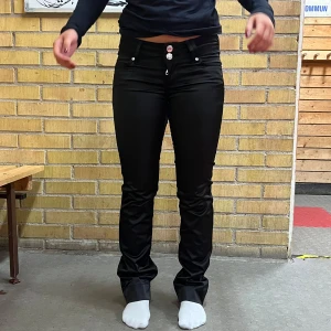 Svarta jeansbyxor - Säljer ett par svarta lågmidjade byxor, som är igentligen är straight, men som är långa på mig. De har en klassisk femficksdesign med knappar och dragkedja framtill. Perfekta för både vardag och fest!