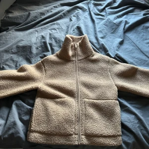 Beige teddyjacka - Mysig beige teddyjacka med dragkedja framtill. Perfekt för kyliga dagar när du vill vara både varm och stilren. Jackan har en hög krage och långa ärmar, vilket gör den extra skön att ha på sig. Den är i ett mjukt och fluffigt material som ger en avslappnad look.