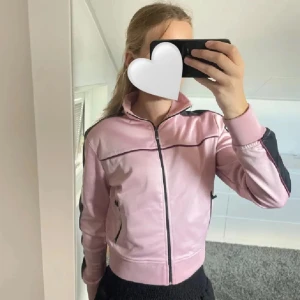 Rosa jacka med svarta detaljer - Snygg rosa jacka med svarta ränder längs ärmarna och dragkedja framtill. Jackan har en hög krage och långa ärmar. Perfekt för en sportig look.