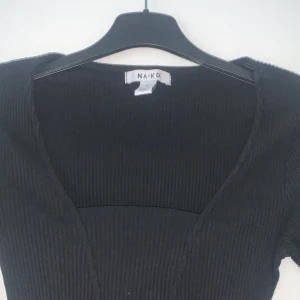 Svart ribbad topp från NA-KD - Snygg svart ribbad topp från NA-KD med lång ärm och djup v-ringning. Perfekt för en stilren look. Toppen är figurnära och passar bra till både jeans och kjol.