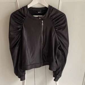 Tunn jacka från Gina Tricot - Tunn och mjuk svart jacka från Gina Tricot med dragkedja framtill. Jackan har långa ärmar med rynkade detaljer vid axlarna och är i ett material som ser ut som skinn. Storlek 44 men upplevs mer som 38/40. 