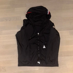 Svart vindjacka från Moncler - Snygg svart vindjacka från Moncler med huva och dragkedja. Jackan har Monclers logotyp på ärmen och en stilren design. Perfekt för blåsiga dagar. Small=1