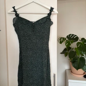 Mörkgrön klänning med vita hjärtan - Charmig mörkgrön klänning med små vita hjärtan och smala axelband. Perfekt för en somrig look med sin lätta och luftiga design. Klänningen har en söt detalj framtill och en smickrande passform.