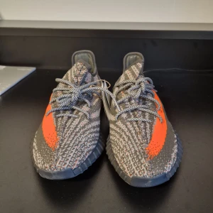 YEEZY BOOST 350 V2  - Snygga Adidas Yeezy Boost 350 V2 med en iögonfallande grå och orange färgkombination. Skorna har en stickad ovandel med snörning och den ikoniska SPLY-350 texten på sidan. Sulan är designad för komfort och stil. Har order bekräftelse från Adidas för att bevisa att de är äkta.