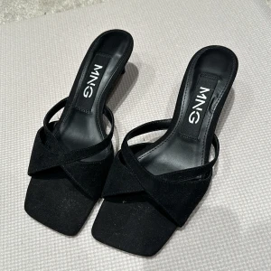 Svarta klackskor från MANGO - Eleganta svarta klackskor från MANGO med öppen tå och korsade remmar. Skorna har en stilren design med en låg klack, perfekt för en chic look. Tillverkade i äkta mocka som ger en lyxig känsla.