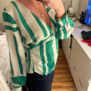 Grön och vit randig blus från Vero Moda - Säljer en snygg grön och vit randig blus från Vero Moda. Blusen har en omlott-design med långa ärmar och en v-ringad hals. Perfekt för en stilren look.