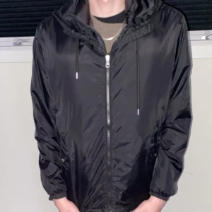 Svart vindjacka från Moncler - Snygg svart vindjacka från Moncler med huva och dragkedja framtill. Jackan har långa ärmar och Moncler-logga på ärmen. Den har också en cool randig detalj i rött, vitt och blått på huvan. Perfekt för en stilren look.