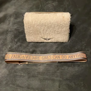 Beige axelväska från Zadig & Voltaire - Säljer en snygg beige axelväska från Zadig & Voltaire med fluffigt yttre och präglad logga. Väskan har en justerbar axelrem med text och flera fack inuti för smart förvaring. Perfekt för att ge din outfit en trendig touch.