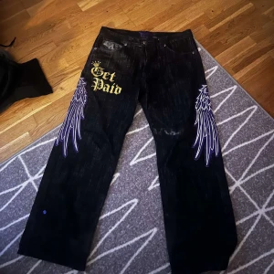 Svarta jeans med tryck - Unika svarta jeans med guldtexten 'Get Paid' och detaljerade lila vingar på benen. Baktill finns ett tryck med en kungakrona och sedlar. Perfekt för dig som vill sticka ut med en cool och edgy stil.