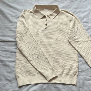 Beige pikétröja från John Henric - Säljer en stilren beige pikétröja från John Henric. Tröjan har en texturerad yta med tre knappar framtill och en klassisk krage. Perfekt för en avslappnad men ändå elegant look.