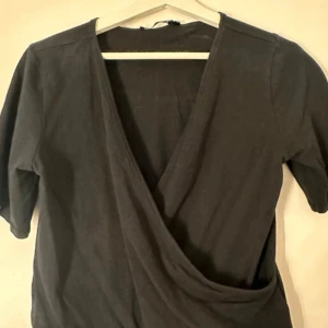 Svart omlottopp från 157 - Snygg svart omlottopp från 157 i storlek M. Toppen har trekvartsärmar och en stilren design som passar perfekt till olika tillfällen. Perfekt för dig som gillar en enkel men elegant stil.