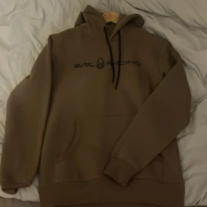 Beige hoodie från Sail Racing - Säljer en beige hoodie från Sail Racing med svart logotyp på bröstet. Tröjan har en klassisk känguruficka och justerbar huva med svarta snören. Perfekt för en avslappnad stil.
