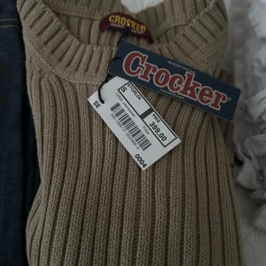 Beige stickad tröja från Crocker - Säljer en beige stickad tröja från Crocker. Tröjan har en ribbad design med förstärkta axelpartier och en klassisk rund hals. Perfekt för kyligare dagar och ger en stilren look.