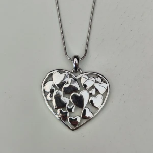Halsband med hjärthänge från Glitter - Elegant halsband med ett stort hjärthänge i silverfärg från Glitter. Hänget har ett vackert mönster av flera små hjärtan. Perfekt för att ge en romantisk touch till din outfit. Inte äkta.