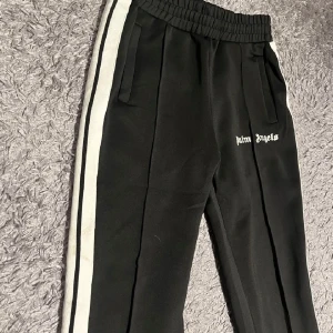 Svarta byxor från Palm Angels - Snygga svarta byxor från Palm Angels med vita sidoremsor och elastisk midja. Byxorna har en avslappnad passform och är perfekta för en trendig look. Logotypen är tryckt på sidan.