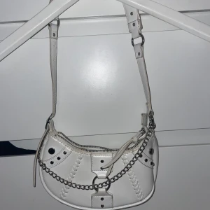 Vit axelväska med kedjedetalj - Snygg vit axelväska med coola kedjedetaljer och nitar. Väskan har en dragkedja upptill och justerbar axelrem. Perfekt för att ge din outfit en edgy touch.