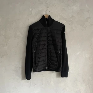 Svart quiltad jacka från Moncler - Snygg svart quiltad jacka från Moncler med dragkedja framtill. Jackan har långa ärmar och en stilren design som passar perfekt för kyligare dagar. Den har en diskret logga på ärmen och ribbade muddar för extra komfort.