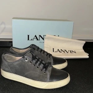Grå sneakers från Lanvin - Snygga grå sneakers från Lanvin med svarta detaljer och snörning. Skorna har en vit sula och är tillverkade i skinn och mocka. Perfekta för en stilren look.