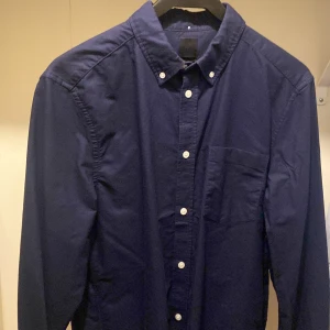 Mörkblå skjorta med knappar - Snygg mörkblå skjorta med klassisk krage och knappar hela vägen ner. Skjortan har en bröstficka och långa ärmar, perfekt för en stilren look. Passar bra till både jeans och chinos.