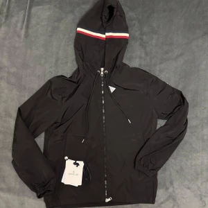 Moncler grimpeurs windbreaker - Snygg svart vindjacka från Moncler med huva och dragkedja. Jackan har en röd och vit rand på huvan och Moncler-logga på ärmen. Perfekt för blåsiga dagar. Använd ett par gånger. Priset kan diskuteras