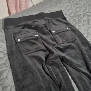 Svarta mjukisbyxor från Juicy Couture - Snygga svarta mjukisbyxor från Juicy Couture med bred resår och snörning i midjan. Byxorna har fickor med knappar bak och en mjuk, glansig yta som ger en lyxig känsla. Perfekta för en avslappnad stil. I väldigt fin skick använd fåtal gångar i storlek S. 