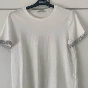 Vit t-shirt med glittriga detaljer från Zara - Snygg vit t-shirt från Zara med glittriga detaljer på ärmsluten. Perfekt för att lägga till lite bling i vardagen. Den har en klassisk rund hals och korta ärmar.