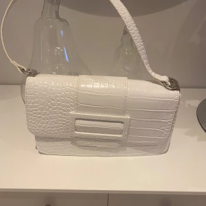 Vit axelväska med krokodilmönster - Snygg vit axelväska med krokodilmönster och stilren design. Väskan har en praktisk axelrem och stängs med magnetknappar. Perfekt för att ge en elegant touch till din outfit.