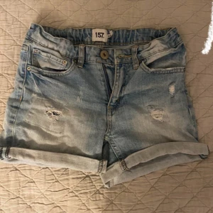 Ljusblå jeansshorts från 157 - Snygga ljusblå jeansshorts från 157 med slitna detaljer och uppvikta ben. Perfekta för en avslappnad stil. De har klassisk femficksdesign och knappgylf.