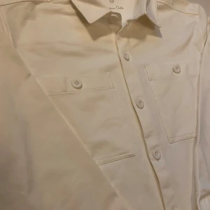 Vit overshirt från Massimo Dutti - Snygg vit overshirt från Massimo Dutti med långa ärmar och knappar framtill. Skjortan har två bröstfickor med knappar och en klassisk krage. Perfekt för en stilren look. Aldrig använd.