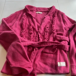 Rosa stickad kofta från Odd Molly - Säljer en snygg rosa stickad kofta från Odd Molly med unika detaljer och broderi på ärmen. Koftan har ett bälte i midjan och dekorativa knappar framtill. Perfekt för att ge din outfit en färgklick!