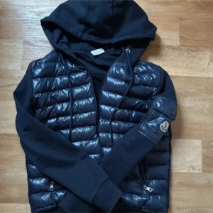 Svart jacka med huva från Moncler - Snygg svart jacka från Moncler med huva och dragkedja. Jackan har en quiltad design på framsidan och ärmar i ett mjukare material. Perfekt för en stilren look.