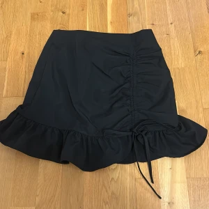 Svart kjol med volang från Shein - Snygg svart kjol från Shein med volangdetalj och knytning i sidan. Perfekt för en stilren look. Kjolen har en kort längd och är tillverkad i ett lätt material.