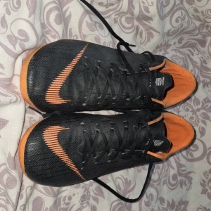 Svarta och orange Nike Mercurial fotbollsskor - Säljer ett par svarta och orange Nike Mercurial fotbollsskor med snörning. Skorna har en sportig design med den ikoniska Nike-loggan i orange på sidan. Perfekta för fotbollsplanen!