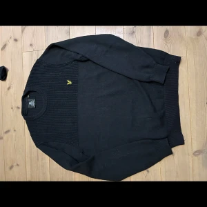 Svart stickad tröja från Lyle & Scott - Säljer en stilren svart stickad tröja från Lyle & Scott med deras ikoniska logga på bröstet. Tröjan har långa ärmar och ribbade muddar vid ärmslut och nederkant. Perfekt för kyligare dagar.