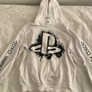 Vit hoodie med PlayStation-tryck från H&M - Säljer en vit hoodie från H&M med stort PlayStation-tryck i svart på framsidan. Hoodien har långa ärmar med PlayStation-logga och text på ärmarna. Perfekt för gaming-entusiaster! 🎮