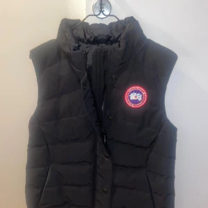 Svart dunväst från Canada Goose - Svart dunväst från Canada Goose. Västen har en dragkedja och fickor framtill och är perfekt för kyligare väder. Den är stilren och funktionell, idealisk för lager-på-lager under vår och höst.