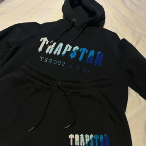 Trapstar Tracksuit - Säljer en svart Tracksuit från Trapstar med texten 'It's a Secret' i blå och vit gradient på bröstet. Hoodien har en klassisk design med dragsko i huvan och är tillverkad i en bomulls- och polyesterblandning. Perfekt för en avslappnad stil.