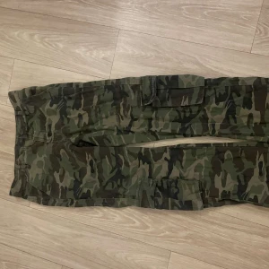 Camouflage cargobyxor - Säljer ett par coola cargobyxor i klassiskt grönt camouflagemönster. Byxorna har flera praktiska fickor och en loose passform som ger en avslappnad stil. Perfekta för en casual look.