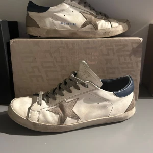Sneakers från Golden Goose - Snygga sneakers från Golden Goose i beige med grå stjärndetaljer och mörkblå häl. Skorna har en vintage-look med snörning och perforerade detaljer på tungan. Perfekta för en avslappnad stil. Storlek 44. Skick 7/10 defekt under skon som är jätte vanligt med dessa skor. Går att fixa. Hör av dig om du undrar något👍