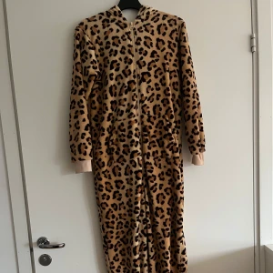 Leopardmönstrad onesie med dragkedja - Mysig och bekväm onesie i leopardmönster med dragkedja framtill. Perfekt för att hålla sig varm och stilren hemma. Den har långa ärmar och en huva för extra komfort.