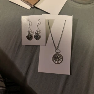 Silverfärgat smyckeset med halsband och örhängen - Elegant smyckeset i silverfärg med ett halsband och matchande örhängen. Halsbandet har ett runt hänge med ett trädmotiv, medan örhängena har hjärtformade hängen med detaljerade mönster. Perfekt för att ge en stilfull touch till din look.