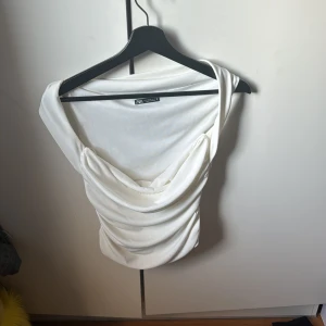 Vit topp från Zara - Säljer en stilren vit topp från Zara med draperad design. Toppen har en elegant offshoulder-stil och är tillverkad i ett mjukt material. Perfekt för en chic look.