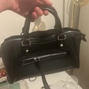 Svart handväska med dragkedja - Snygg svart handväska i skinn med silverfärgade dragkedjor och detaljer. Väskan har dubbla handtag och en praktisk ytterficka med dragkedja. Perfekt för att bära dina dagliga nödvändigheter med stil.  Liknar citybagen och är i väldigt bra skick! Fläckarna man ser på spegelbilden är inte på väskan utan på spegeln! Lämna gärna prisförslag och tveka inte att skriva om du undrar något! 💗