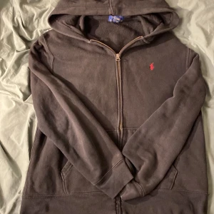 Svart hoodie från Polo Ralph Lauren - Säljer en klassisk svart hoodie från Polo Ralph Lauren med dragkedja och röd broderad logga på bröstet. Perfekt för en avslappnad stil med praktiska fickor och huva. Passar både till vardags och chill.