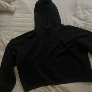 Svart croppad hoodie - Säljer en svart croppad hoodie med luva och dragsko. Perfekt för en avslappnad stil. Den har långa ärmar och är gjord i ett mjukt material.