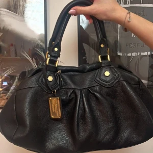 Svart skinnväska från Marc by Marc Jacobs - Snygg svart skinnväska från Marc by Marc Jacobs med guldfärgade detaljer och dragkedja. Väskan har ett stilrent mönstrat innerfoder och en praktisk storlek för alla dina nödvändigheter. Perfekt för att ge din outfit en lyxig touch. Prutat och klart. Skambud=Blokerad.
