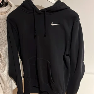 Svart hoodie från Nike - Säljer en klassisk svart hoodie från Nike med den ikoniska loggan på bröstet. Tröjan har en justerbar huva med dragsko och en stor magficka framtill.