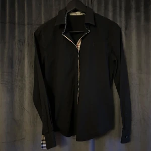 Svart skjorta från Burberry - Snygg svart skjorta från Burberry med klassiskt rutigt mönster på insidan av kragen och ärmsluten.  Dam storlek, finns inte storlek på skjortan men det är en S storlek. Kontakta vid frågor ☺️