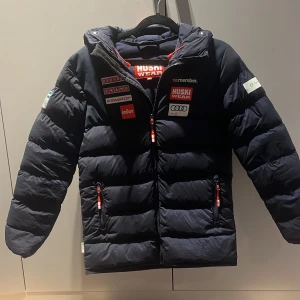 Svart pufferjacka från Huski Wear, limiterad edition  - Säljer en svart pufferjacka från Huski Wear med flera logotyper och märken på framsidan. Jackan har en dragkedja och är långärmad, perfekt för kyliga dagar. Den har en sportig look med röda detaljer på dragkedjorna. Limited edition. Pris kan diskuteras